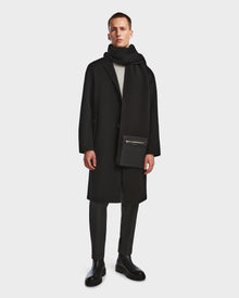 A man stands against a plain background wearing the Rudsak DRAL - 6124503 Black wool jacket styled as a long coat, paired with a black scarf, gray trousers, black boots, and holding a black zippered pouch.