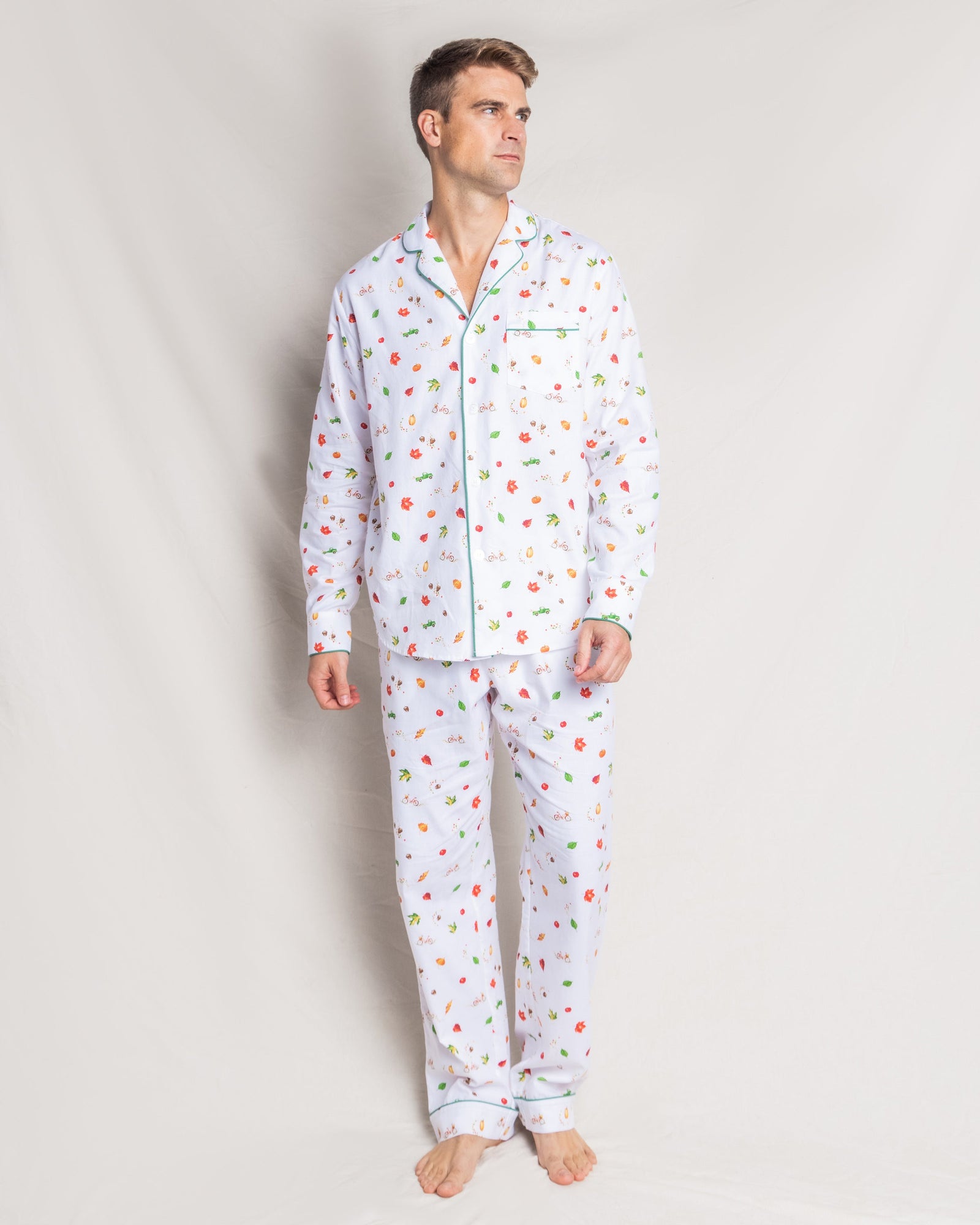 A person stands against a plain background wearing Petite Plumes Mens Twill Pajama Set in Shades of Autumn, featuring a collar, buttons, and a pocket with green piping. Barefoot, the person gazes thoughtfully to the side.