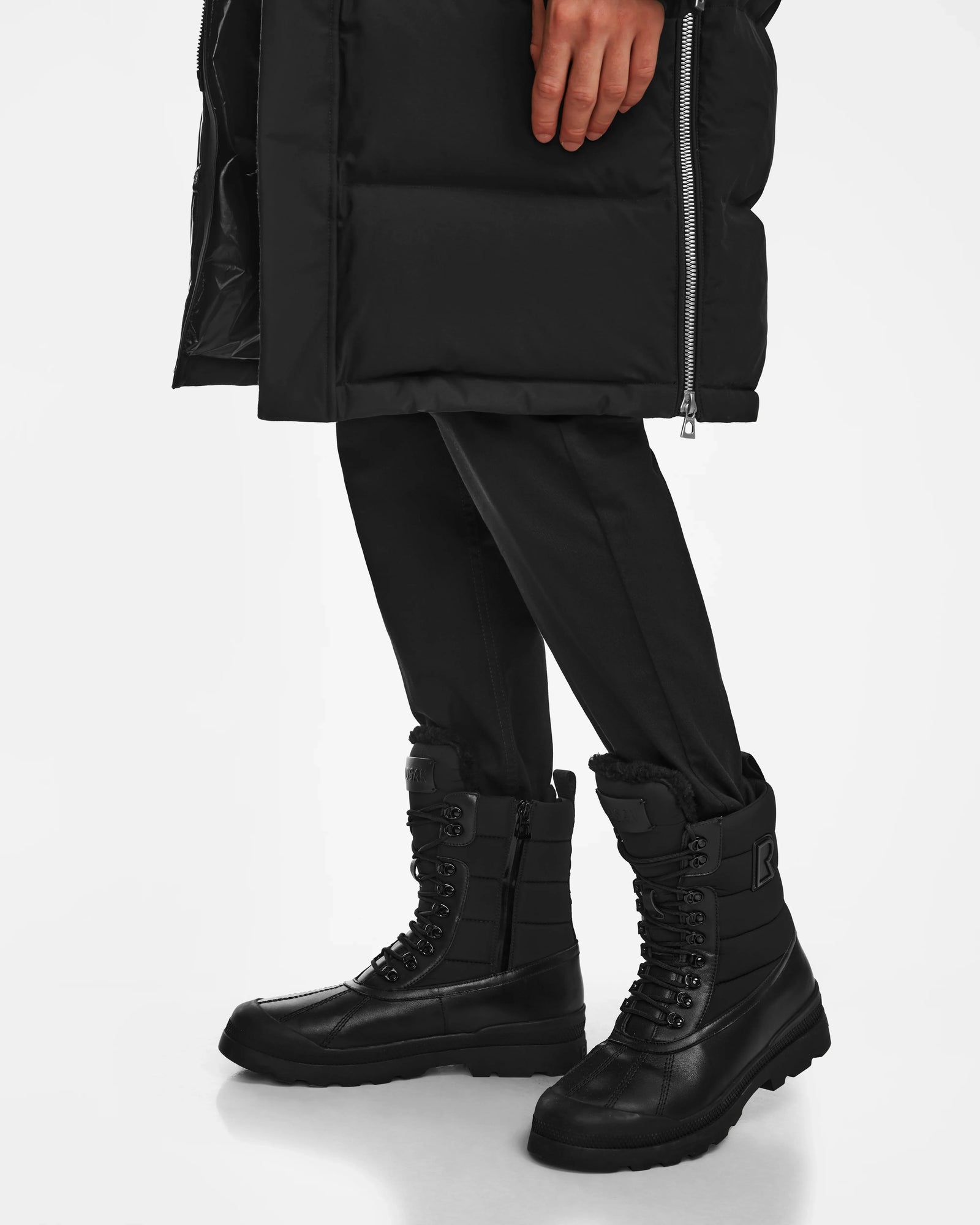 A person wearing Rudsak REAGAN - 6224501 Black men’s winter boots stands on a white background, with only their lower body dressed in black pants and a long black coat visible.