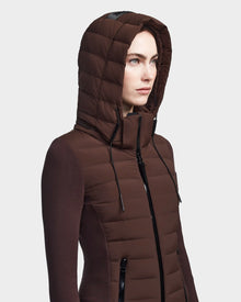 A woman in the RUDSAK MERINA - 8122546 CHOCOLATE jacket, a brown quilted down style with hood up and black zippers, stands against a plain, light background. The sleek women’s jacket has a fitted design.