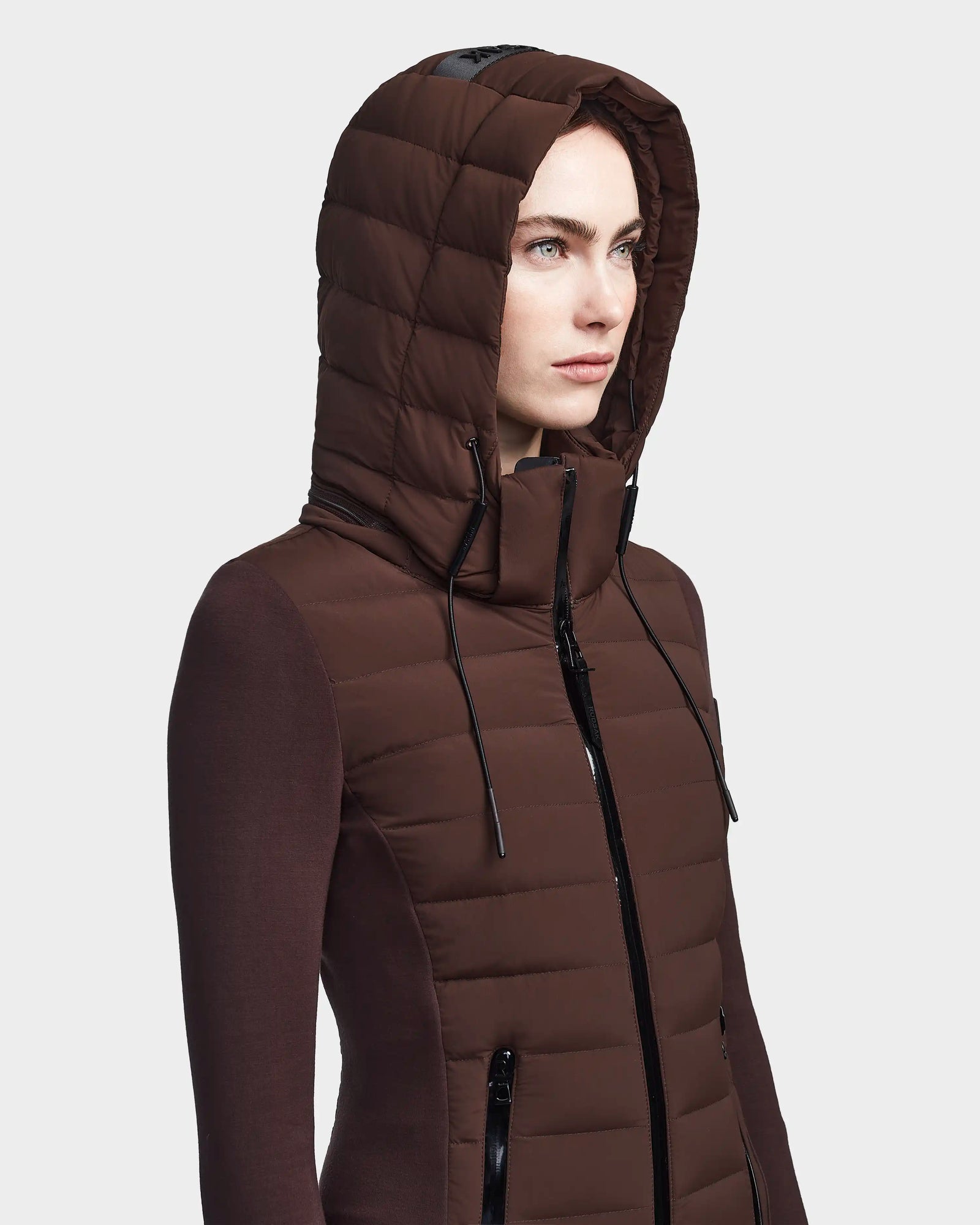 A woman in the RUDSAK MERINA - 8122546 CHOCOLATE jacket, a brown quilted down style with hood up and black zippers, stands against a plain, light background. The sleek women’s jacket has a fitted design.