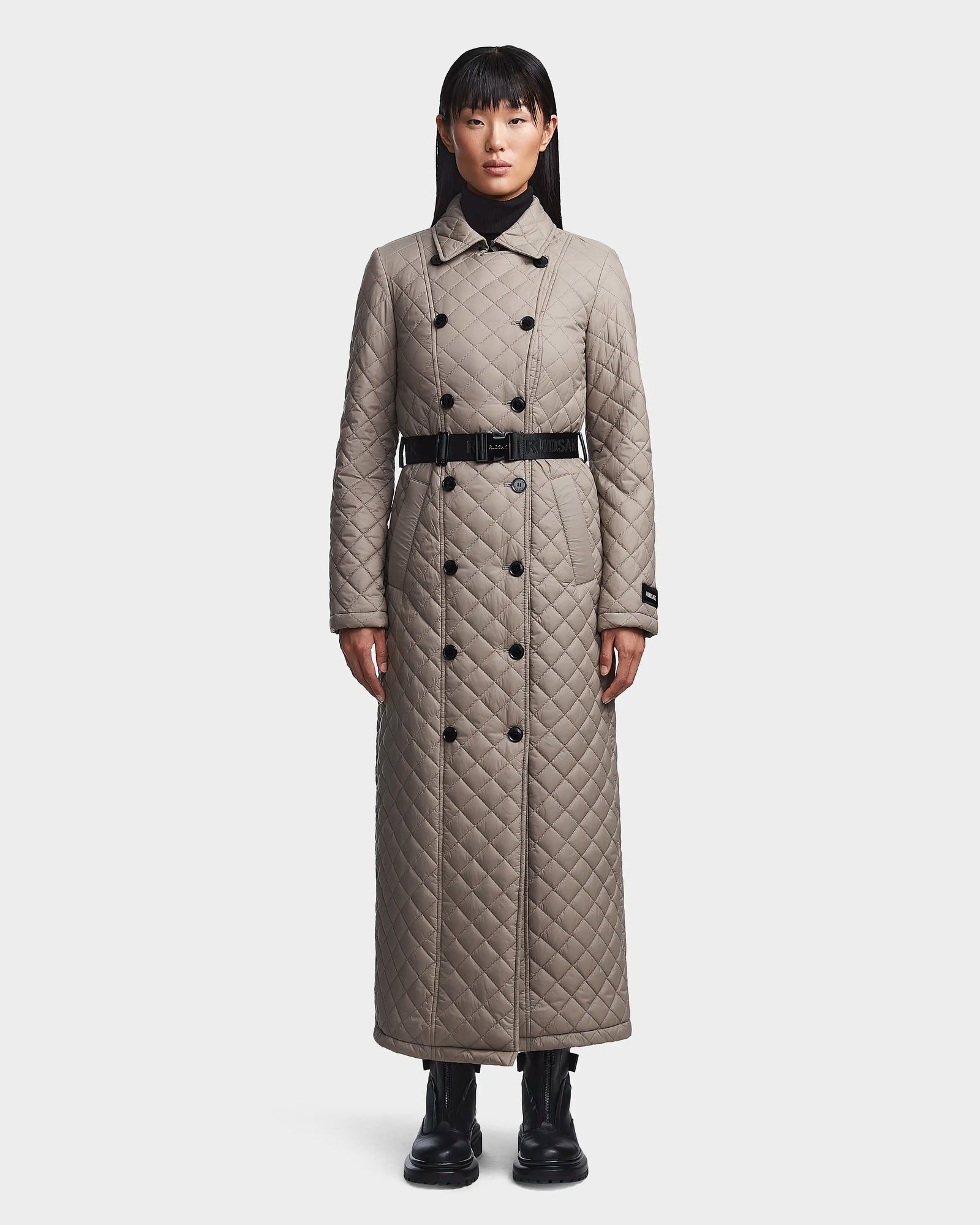 A woman faces forward in the Rudsak DAISY - 8123580 Cement women’s quilted maxi trench coat with sustainable Sorona insulation, double-breasted buttons, black belt, diamond pattern to the ankles, and black boots against a white background.