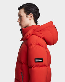 A person with short dark hair in profile wears the Rudsak VINCENT M - 6123510 Orange men's down puffer jacket with a large hood, ultra-warm insulation, signature sleeve patch, and silver zippers against a plain white background.