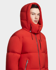 A person stands against a plain light gray background wearing the Rudsak VINCENT M - 6123510 Orange men’s down puffer with ultra-warm insulation and a large hood, partially zipped up.