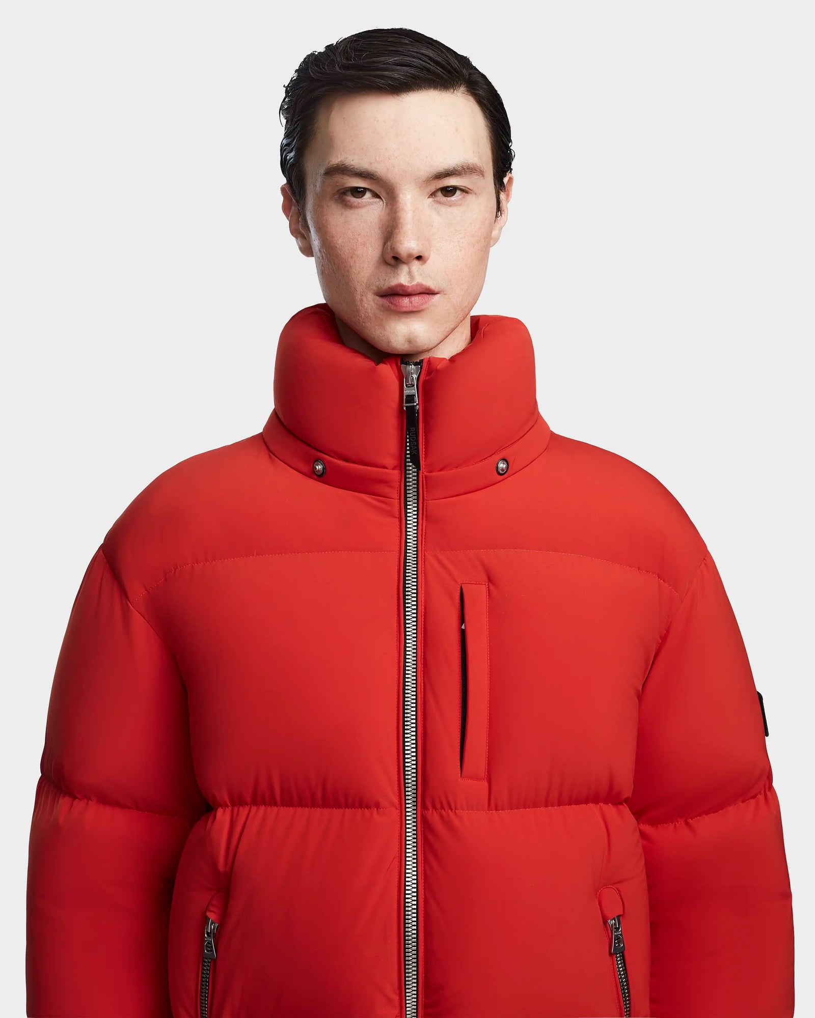 A person with short dark hair wears the Rudsak VINCENT M - 6123510 Orange men's down puffer jacket with a high collar, standing against a plain light gray background.