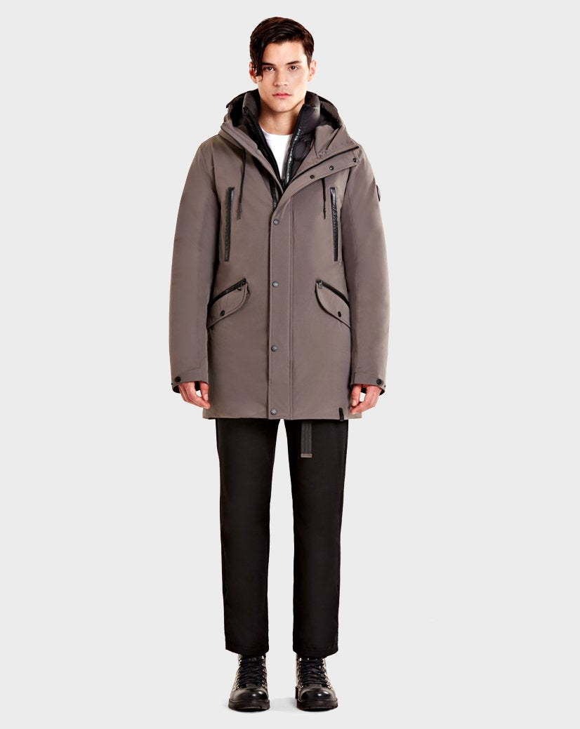 A person stands facing forward, wearing the Rudsak DENT - CA EN 6119515 ELEFANTE parka with leather trim over a white shirt, black pants, and black lace-up boots against a plain light background.