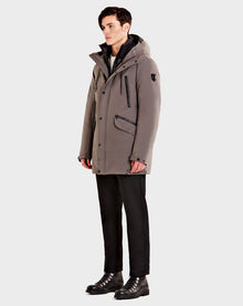 A man stands facing left, wearing the Rudsak DENT - CA EN 6119515 ELEFANTE gray mid-length parka, black pants, and black lace-up boots against a plain light gray background.