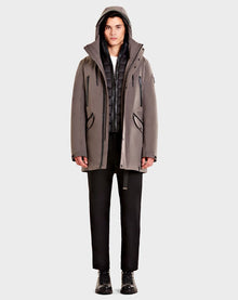 A person faces forward wearing the Rudsak DENT - CA EN 6119515 ELEFANTE gray mid-length parka with leather trim over a black puffer vest, white shirt, black pants, and black boots against a plain light background.