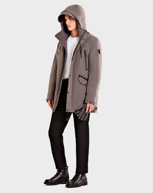 A person wearing the Rudsak DENT - CA EN 6119515 ELEFANTE gray mid-length parka with leather trim, white shirt, black pants, and black boots stands against a plain light background, holding black gloves in one hand.
