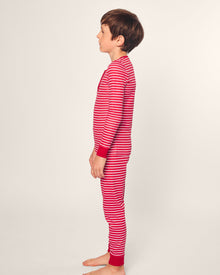 A child stands barefoot in profile against a plain background, wearing Petite Plumes Kids Pima Snug Fit Pajama Set in Red Stripe. The sleepwear features a long-sleeve top and pant set with red and white stripes and red cuffs for a cozy bedtime look.