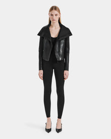 A woman with straight light brown hair and a neutral expression faces forward, wearing the AVRIL - 8124561 Black leather jacket by Rudsak over a black top and fitted pants, paired with black pointed-toe heels.