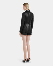 A woman with straight blonde hair wears the Rudsak DANICA - 8125025 Black tailored leather blazer and matching shorts, paired with black heeled sandals, standing against a plain white background.