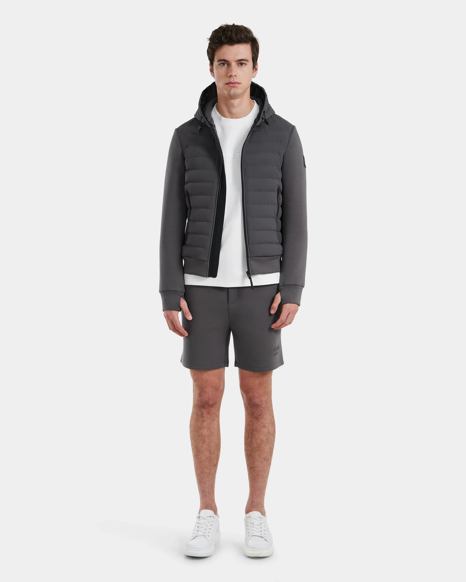 A man faces forward against a plain white background, wearing the Rudsak DEE MAT - 6124028 Grey mens lightweight down jacket, paired with a white t-shirt, gray shorts, and white sneakers.