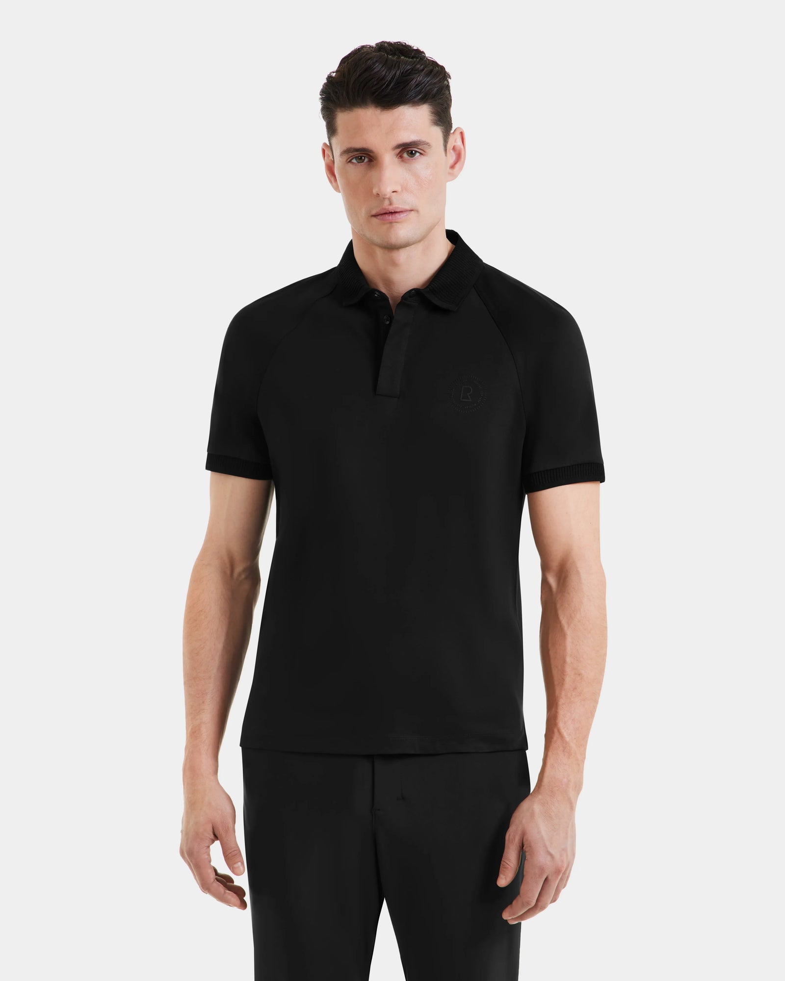 A man with short dark hair wears the Rudsak WALTER - 6524531 Black/Black mens polo and black pants, standing against a plain light gray background.