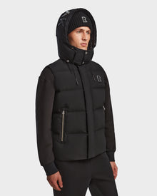A person wears the Rudsak REN M - 9123512 Black M weather-resistant hooded puffer jacket with zip pockets and a black beanie with white embroidered letters, standing against a plain white background.