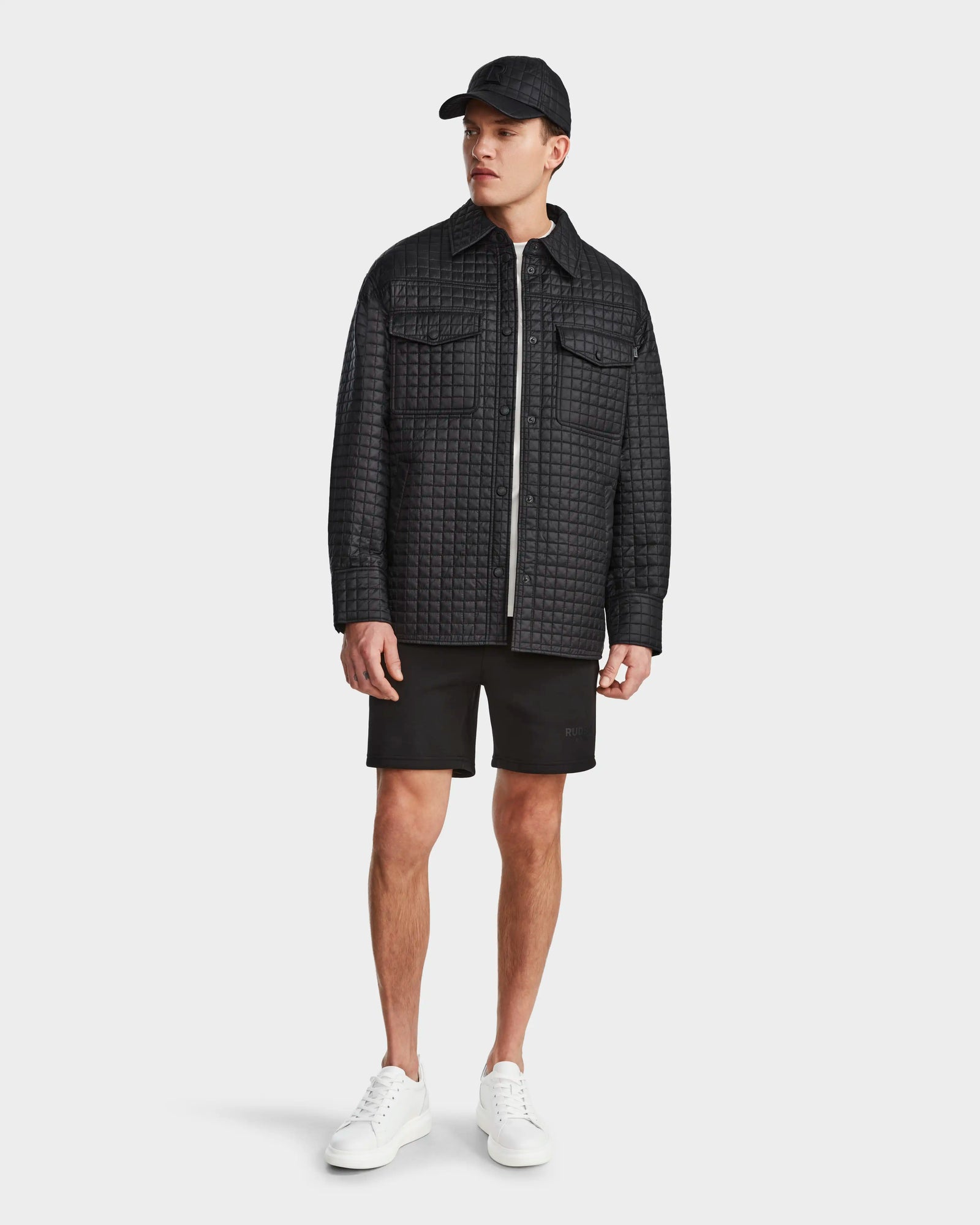 A man in a Rudsak KERRY - 9124003 Black M shirt jacket, paired with black shorts, a black cap, and white sneakers stands against a plain light background, looking to his left.