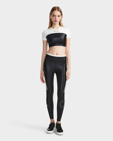 A woman with straight light brown hair stands facing forward, wearing Rudsak’s LILIANE - 8524018 Black women’s fitted crop top and matching high-waisted black leggings with white stripes, paired with black sneakers, against a plain light gray background.