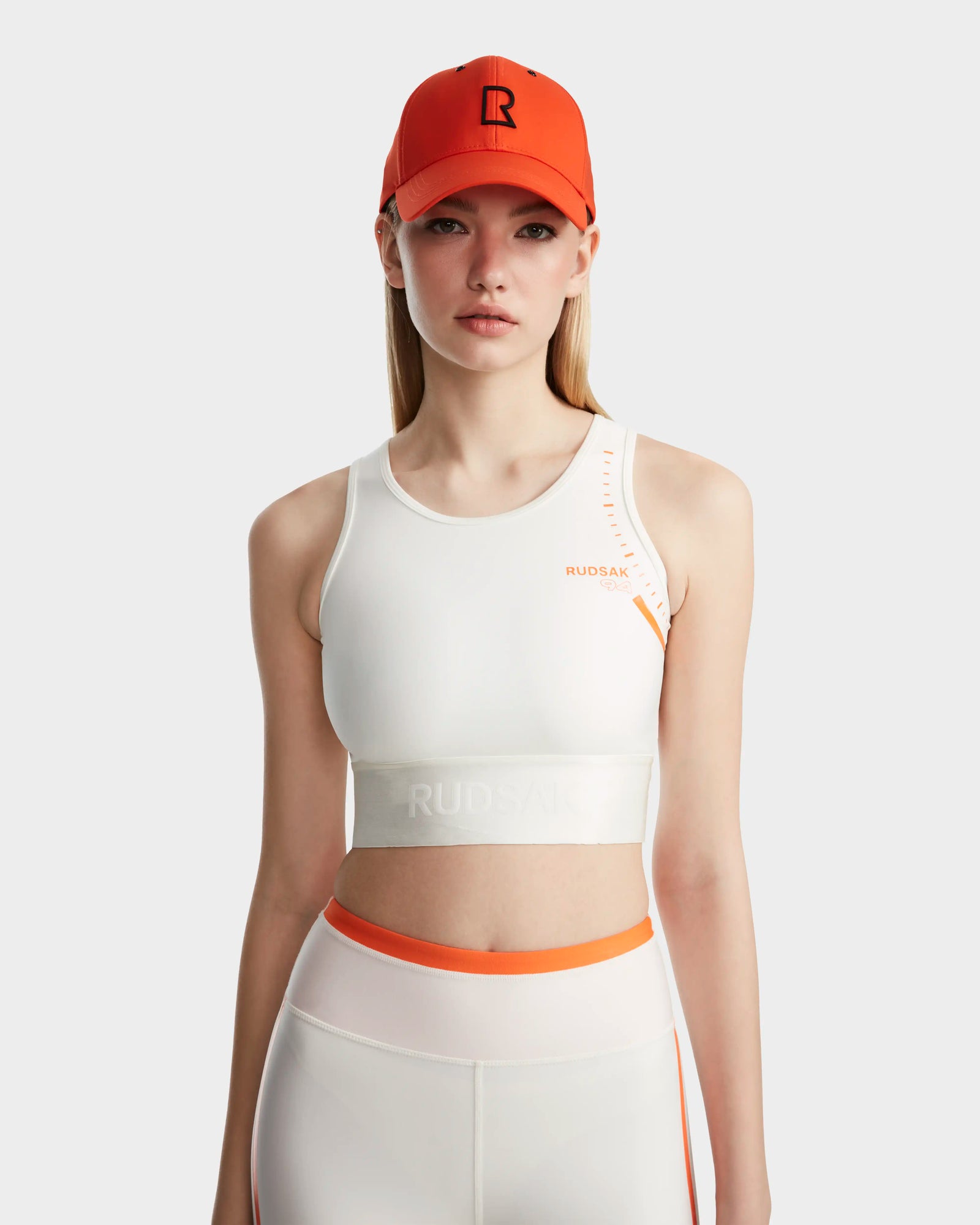 A woman in the Rudsak TRACY - 8524016 Eggshell high-performance sports bra, white crop top, and leggings with orange accents stands against a plain light background, wearing a bright orange baseball cap.
