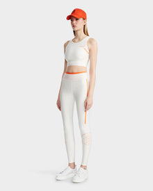 A woman wears the TRACY - 8524016 Eggshell sports bra and leggings by Rudsak, paired with an orange cap and white sneakers, standing against a plain light gray background.