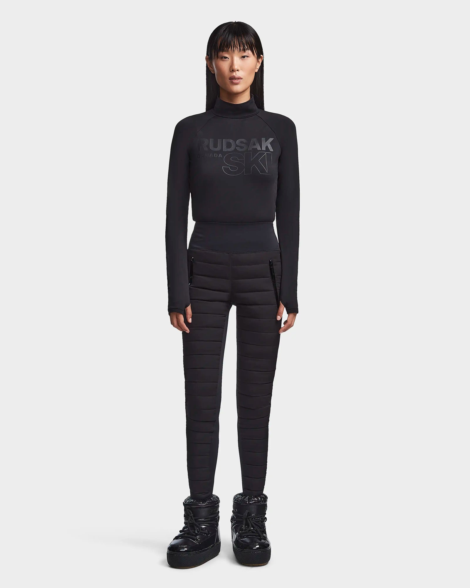 A woman with straight black hair wears the Rudsak KILTON - 8523533 Black fitted top and quilted high-waisted pants with padded boots, creating a chic winter look against a plain white background.