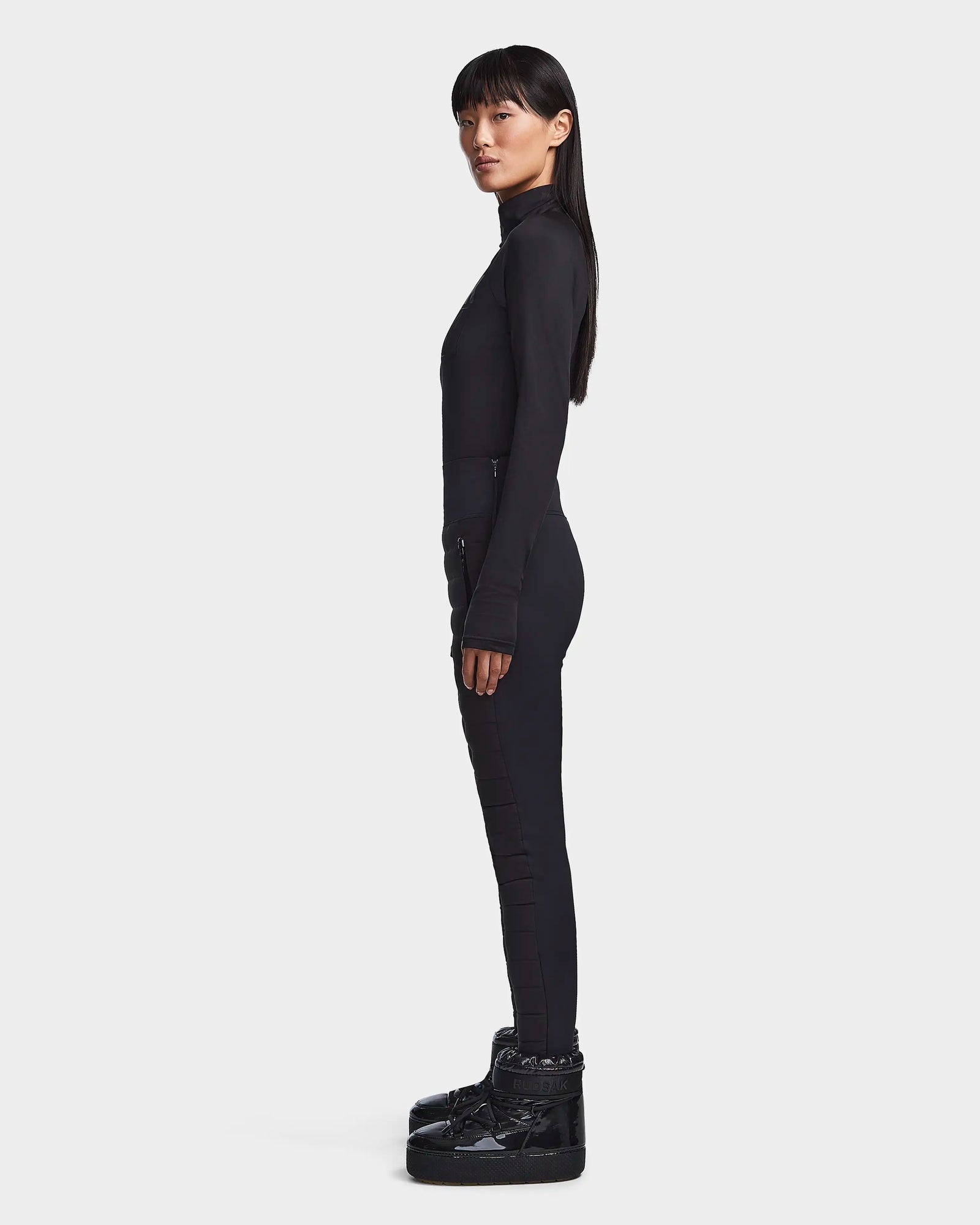 A woman stands in profile against a white background, wearing the Rudsak KILTON - 8523533 Black turtleneck, black pants, and chunky boots—perfect winter staples. Her long, straight hair falls over her shoulders.