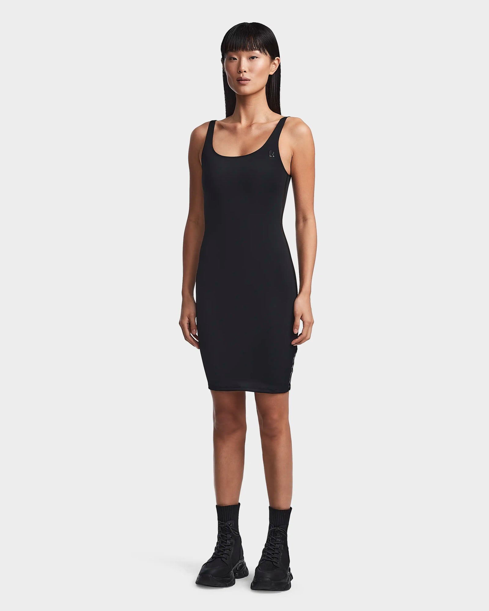 A woman with straight black hair wears the Rudsak MILEY - 8523034 BLACK sleeveless bodycon dress and black combat boots, standing against a plain white background.