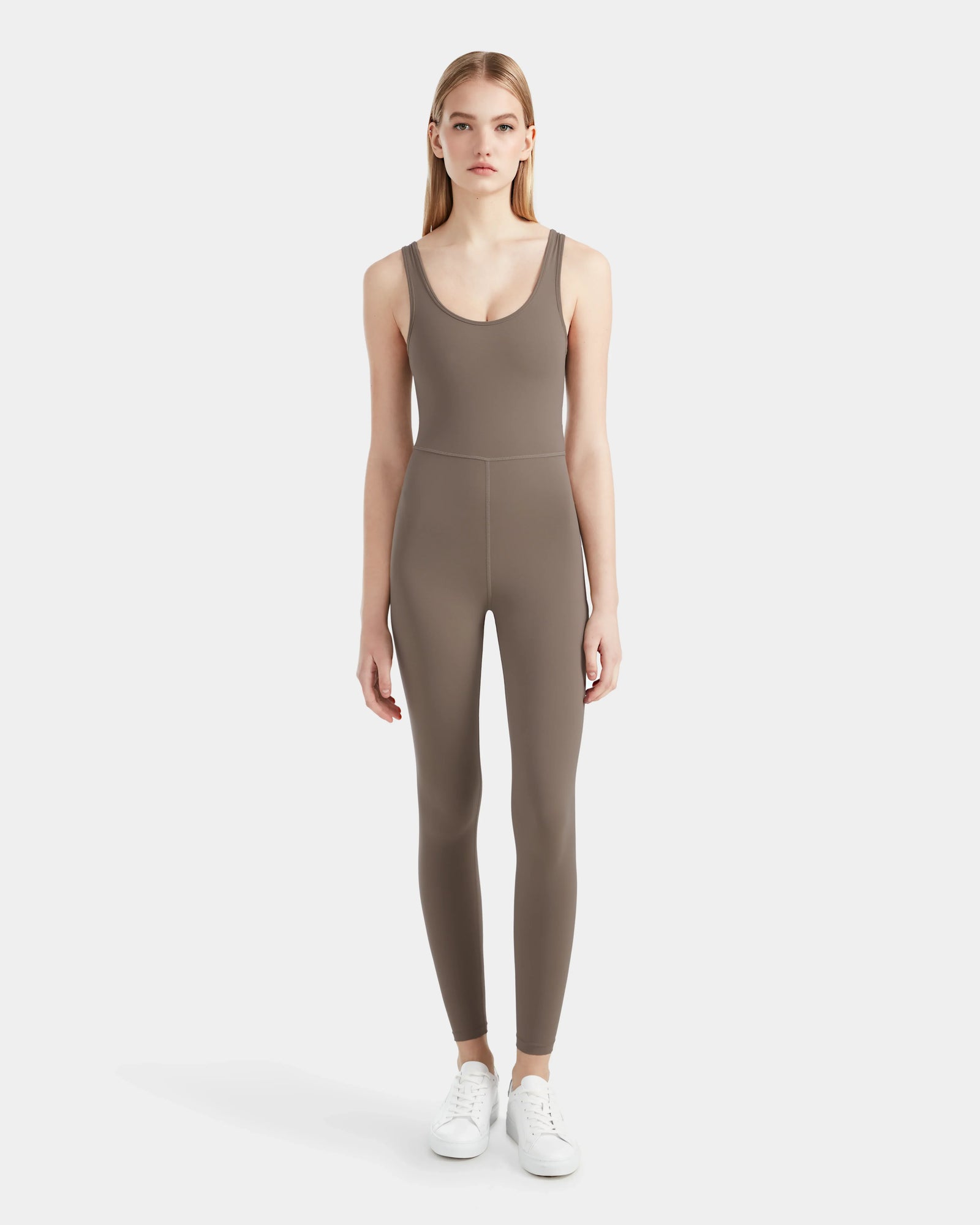 A woman faces forward in the Rudsak JEWEL - 8524032 Cement womens sleeveless stretch jumpsuit, fitted and paired with white sneakers, set against a plain white background.