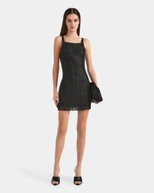 A woman stands facing forward wearing the Rudsak NATALIA - 8524045 Black women's sleeveless, black, grid-patterned mini dress with black heeled sandals. She holds a large black clutch; her long brown hair is straight. The background is plain white.