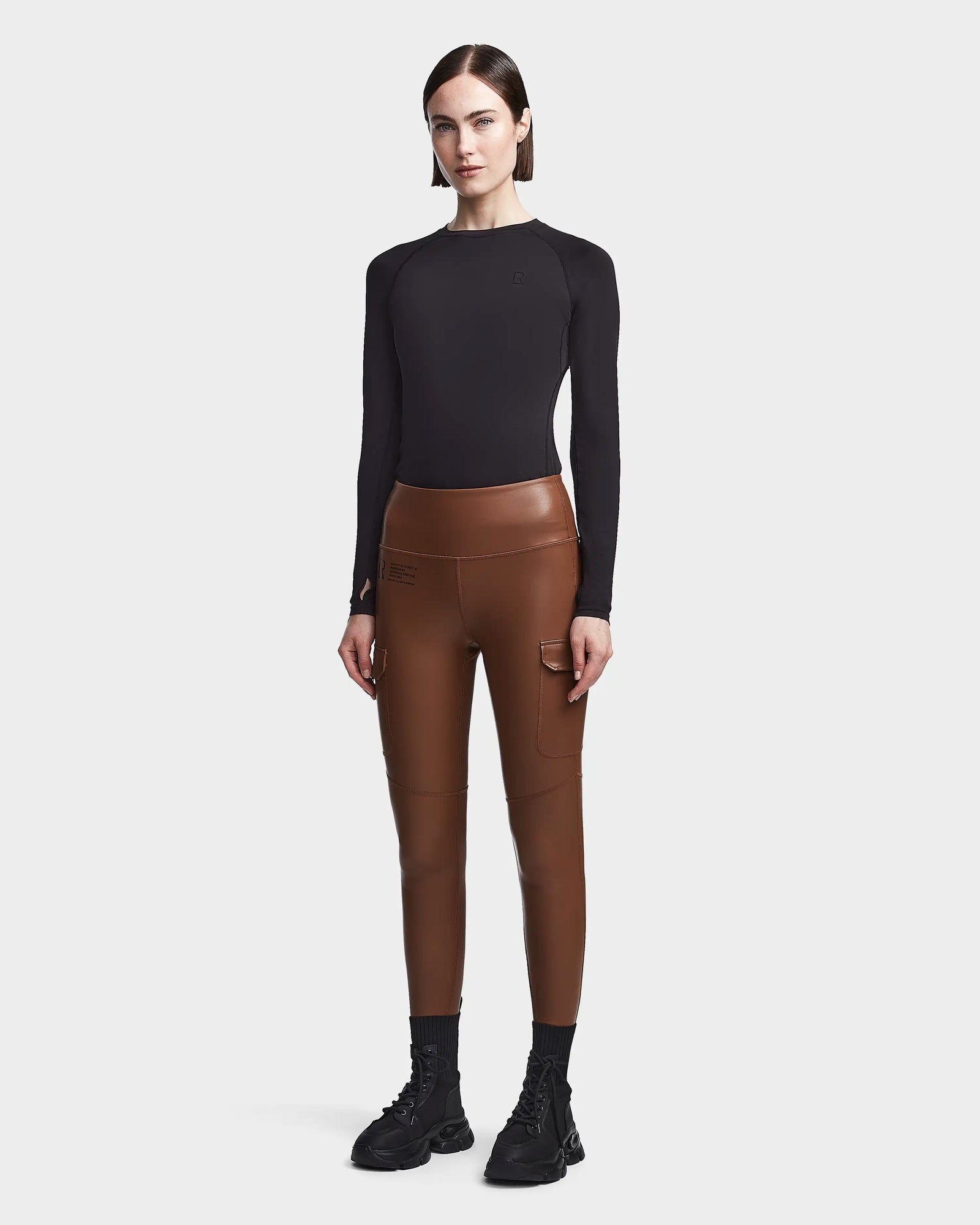 A woman stands against a plain background, wearing Rudsak's SIEN L - 8521042 COHIBA high-waisted brown vegan leather leggings with pockets, paired with a fitted black long-sleeve top, black socks, chunky shoes, and loose straight dark hair.