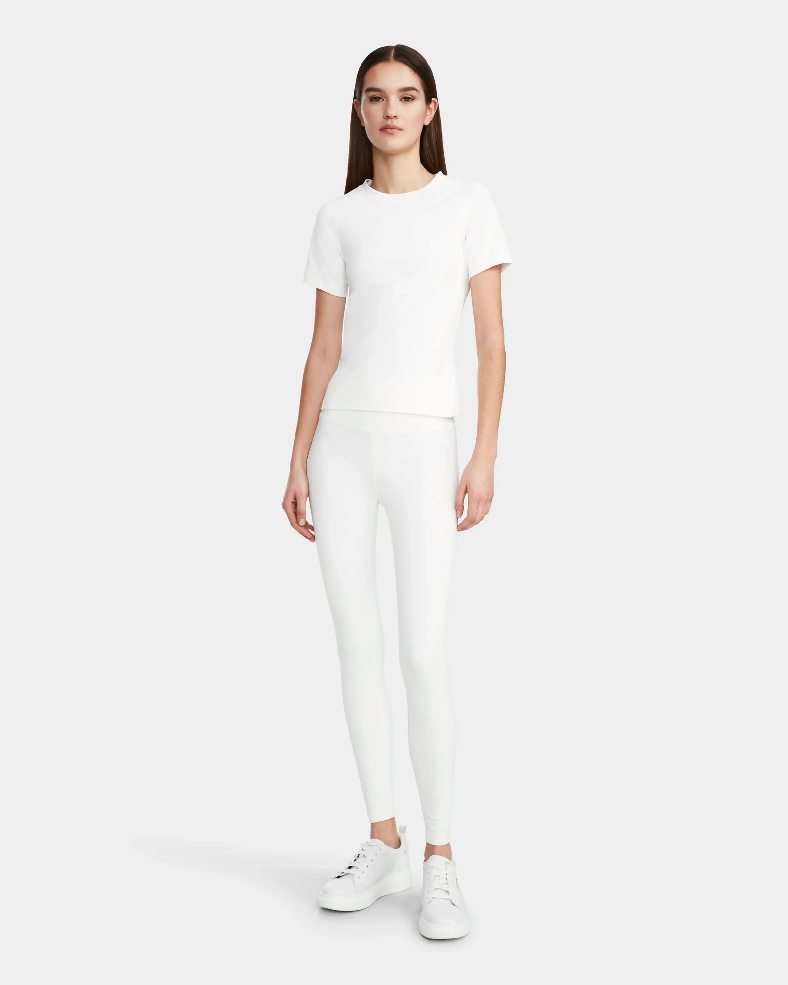 A woman faces forward wearing the Rudsak SUMI - 8524030 Cotton White womens essential tee, paired with white leggings and white sneakers, against a plain light gray background.