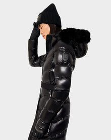 A woman wearing the Rudsak ELSIE - L121525 BLACK shiny black maxi down puffer coat with removable hood, plus a black knit hat and gloves, stands in profile against a plain light background, touching her hat with one hand.