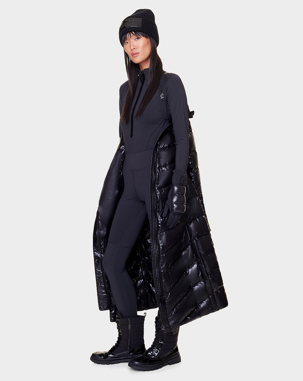 A woman faces forward in a modern, all-black winter look, wearing the Rudsak ELSIE - L121525 BLACK maxi down puffer with a recycled semi-gloss exterior. The stylish coat exudes warmth against a plain background.