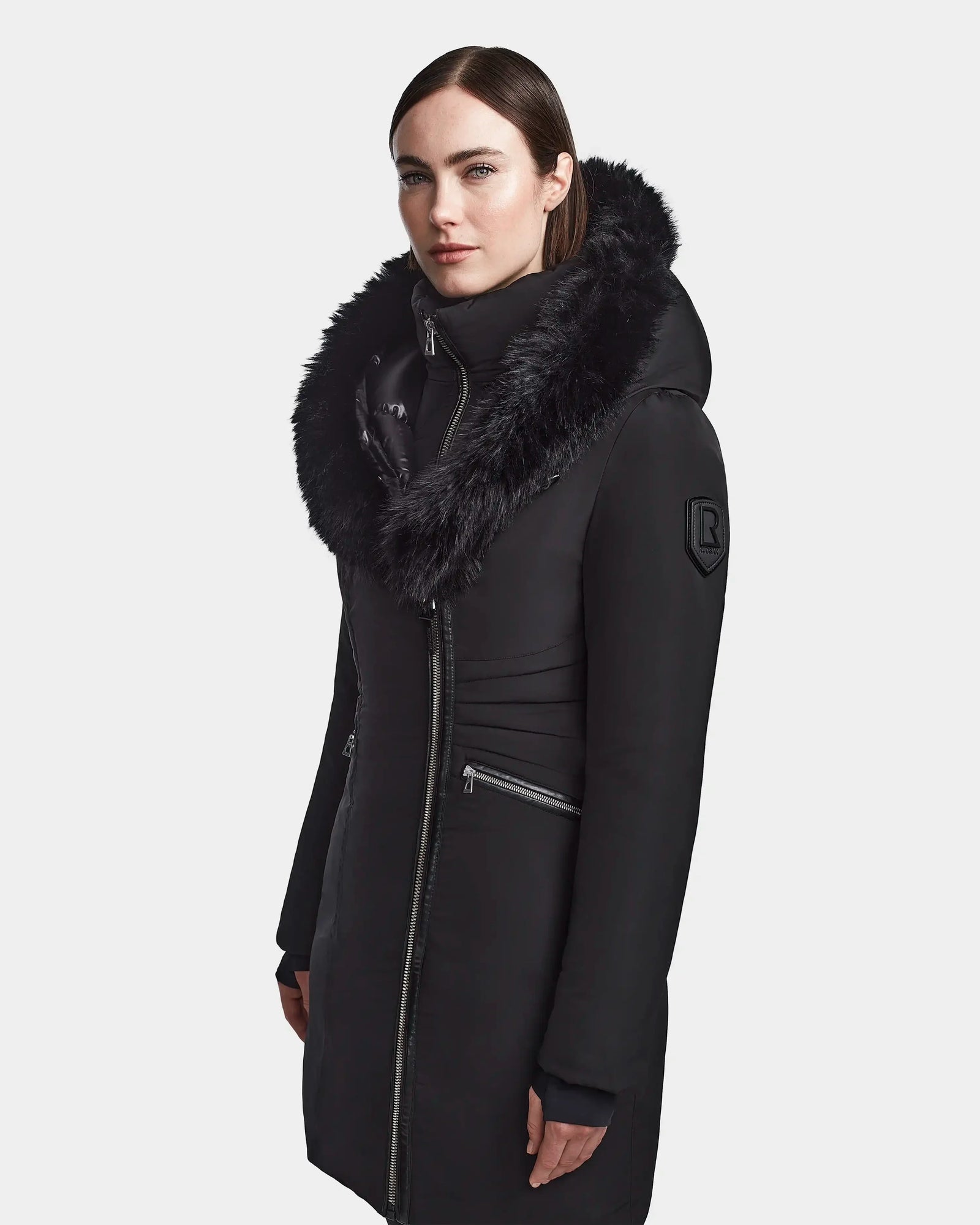 A woman with straight brown hair wears the Rudsak CORA - L123538 Black women's fitted parka, a long black winter coat with a large furry hood and zippered pockets, standing against a plain light background.