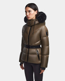 A woman wears the Rudsak CHRISTELLE S - L123524 women's chevron quilted down puffer in light olive, featuring a black fur-trimmed hood, black belt, and zipper details, standing against a plain light gray background.