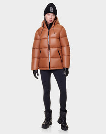 A woman wearing the Rudsak EVIE - L122517 DARK CAMEL puffer, black leggings, gloves, winter boots, and a black beanie with an R stands against a plain white background.