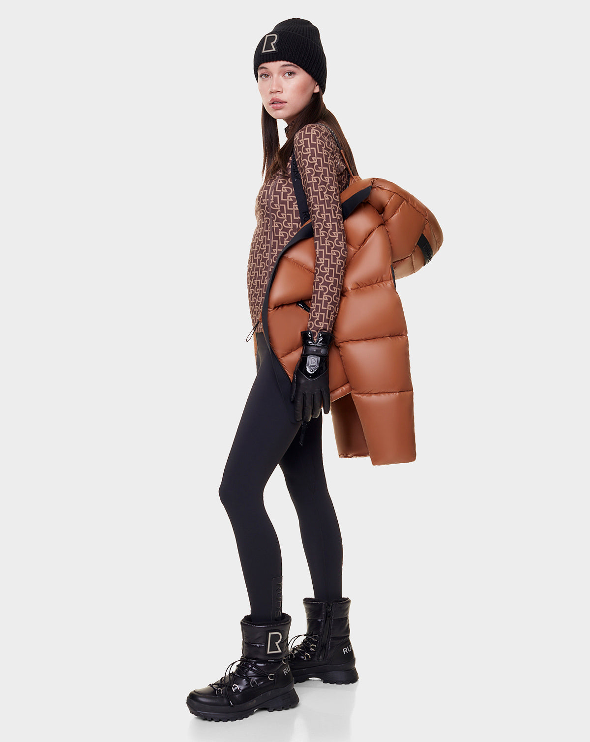 A woman stands sideways against a white background, holding the Rudsak EVIE - L122517 DARK CAMEL RDS-certified down puffer for women over her shoulder. She wears black leggings, a patterned brown top, black boots, gloves, and a beanie.