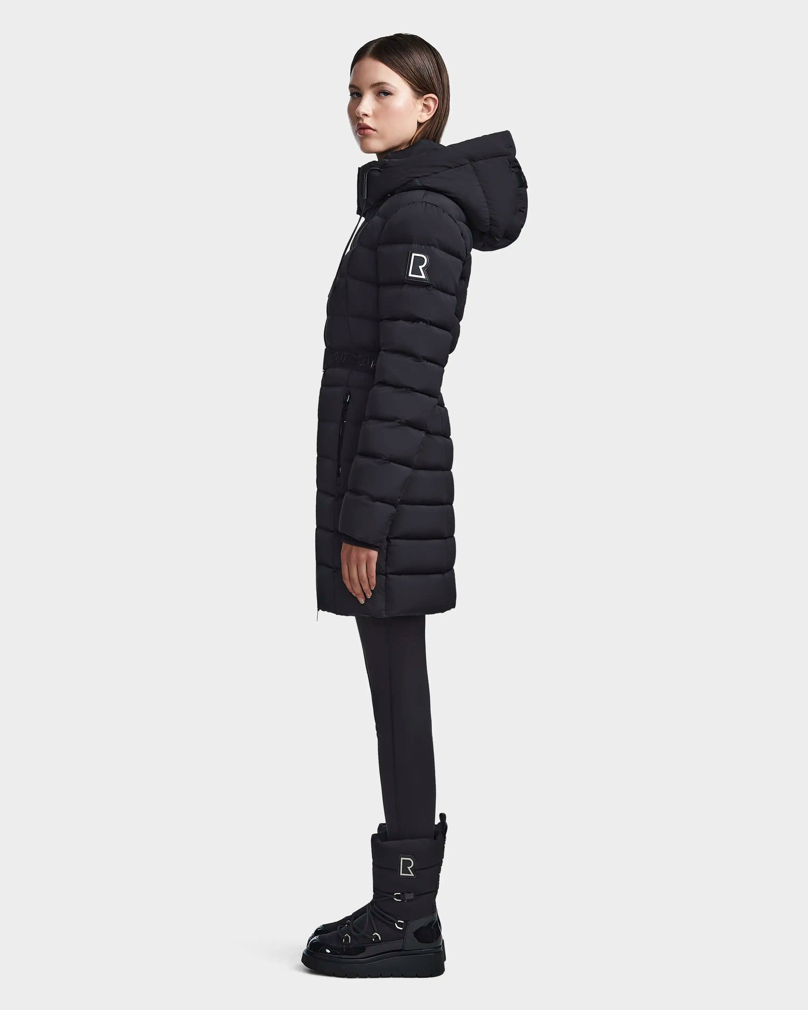 A woman in profile wears the Rudsak FRIDA M - L123514 black mid-length down jacket with removable hood, black pants, and boots. The quilted jacket and boots display a white R logo. The background is plain and light.
