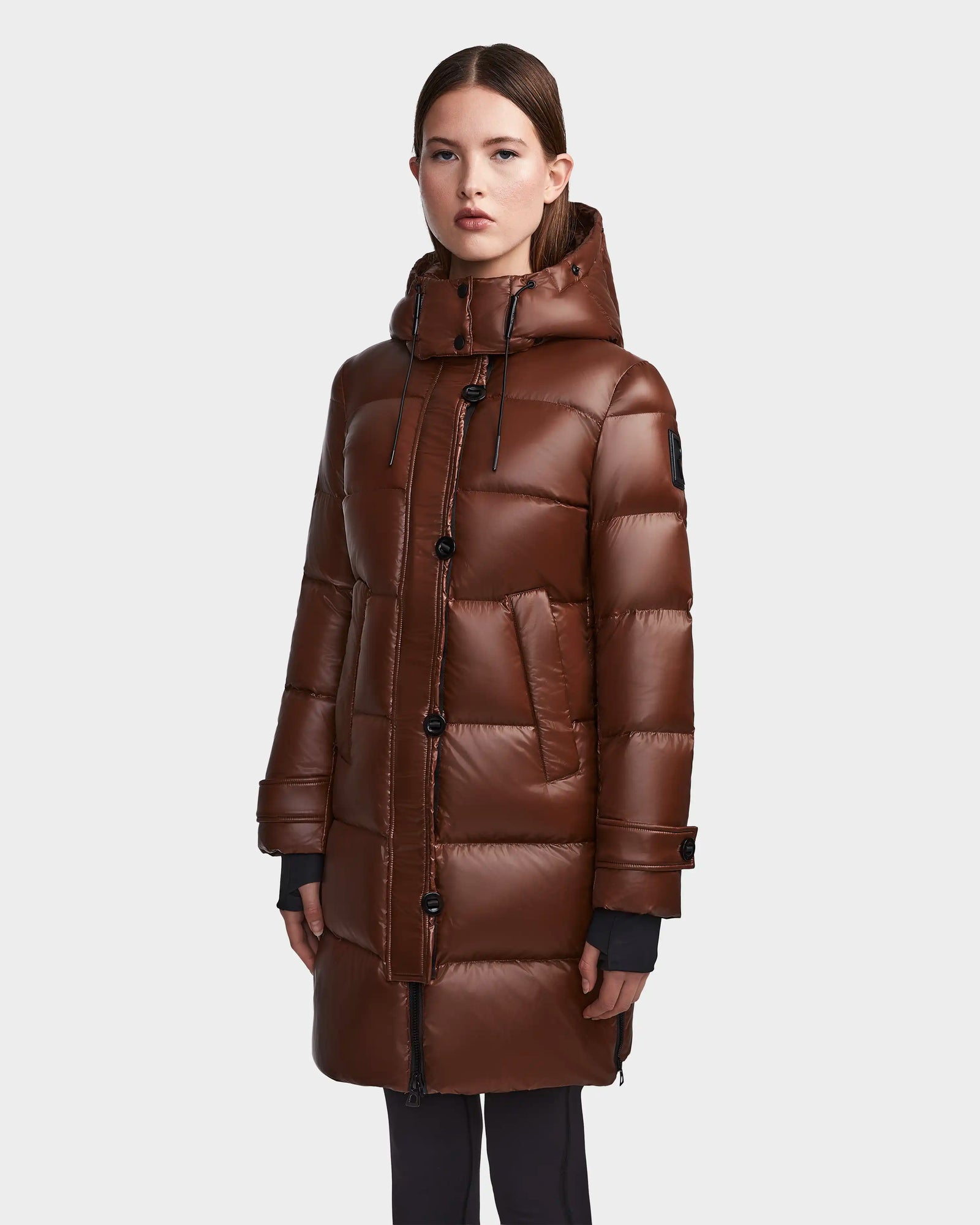 A woman stands facing forward in the Rudsak MEYA S - L123521 Cohiba, a long brown down puffer coat with a shiny quilted finish, black buttons, and a hood. Her arms rest at her sides against a plain background.