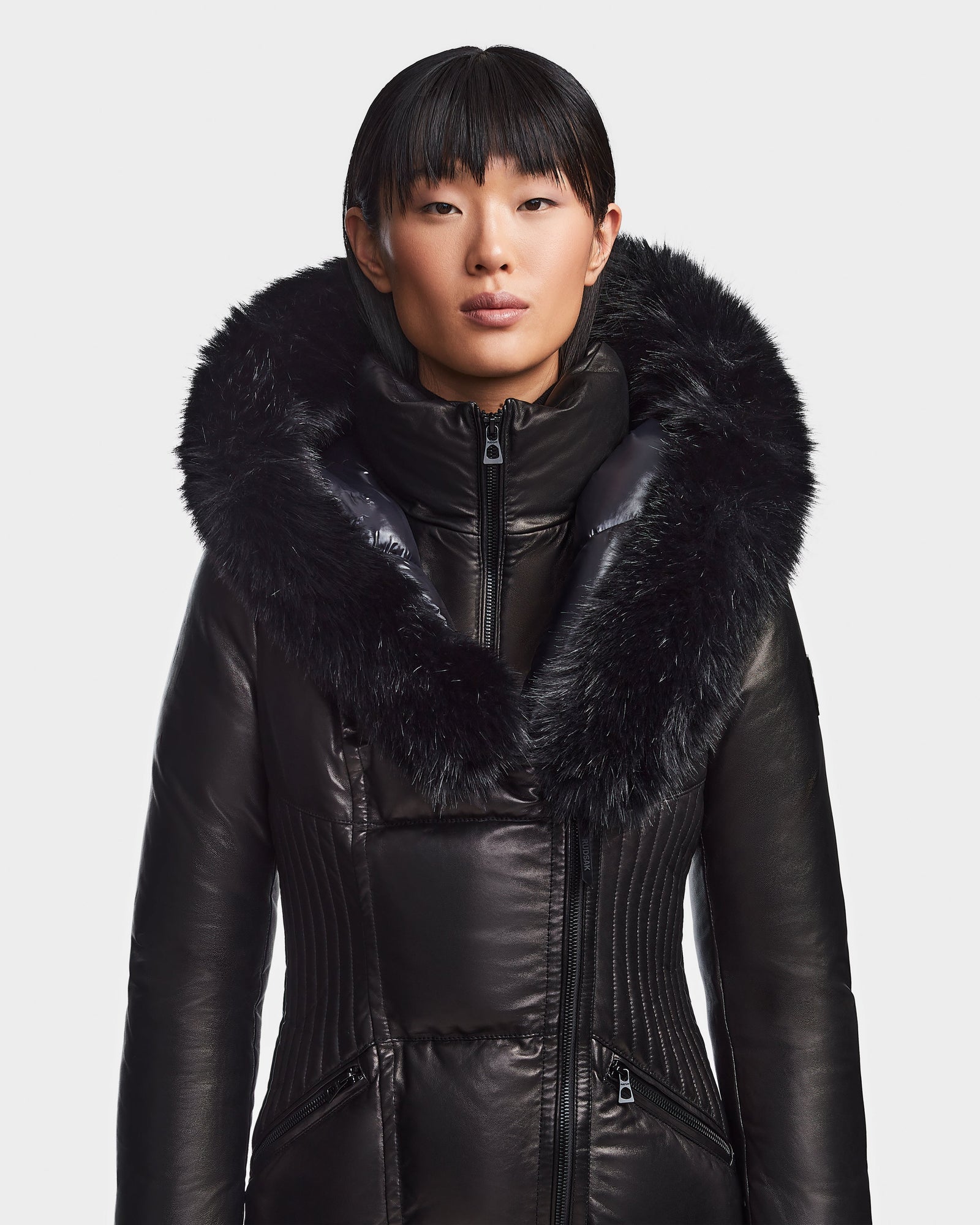 A person wears the SHAUNA X - 8121568 BLACK women's winter coat by Rudsak, featuring cloud leather and a large fur-trimmed hood, standing against a plain white background and looking straight ahead with a neutral expression.
