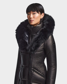 A person wears the Rudsak SHAUNA X - 8121568 BLACK women's winter coat, featuring a sleek design and a large faux fur-trimmed hood, standing against a plain light background.