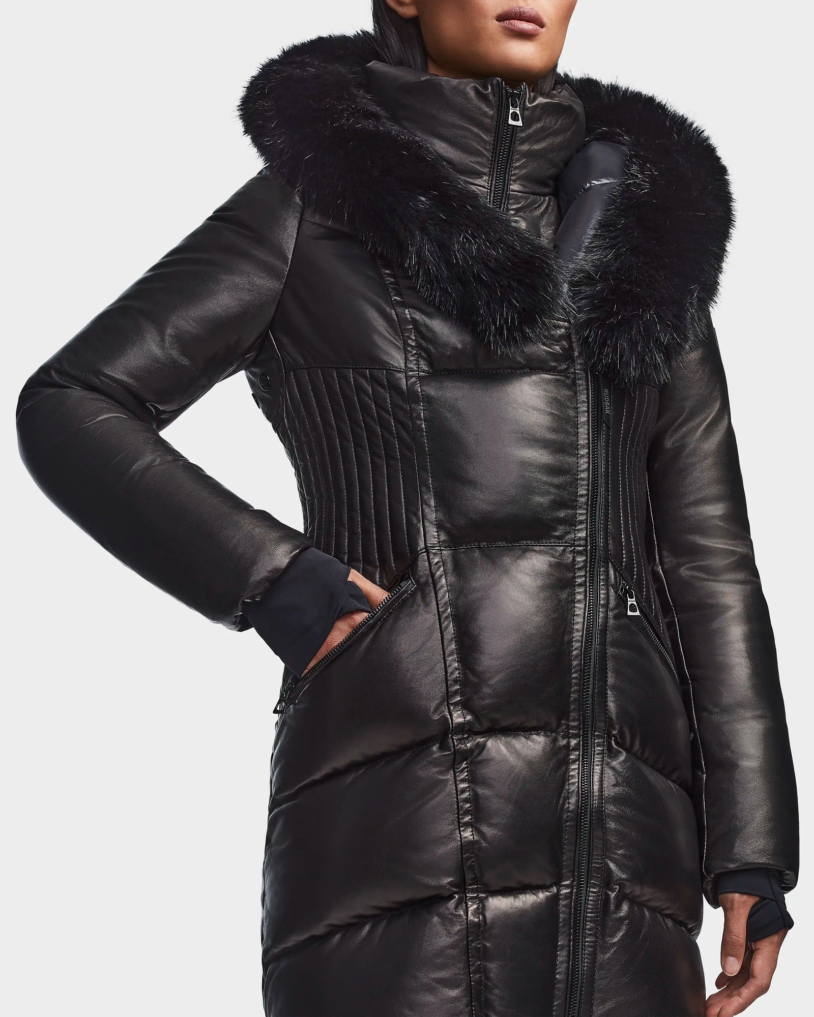 A person wears the Rudsak SHAUNA X - 8121568 BLACK women's winter coat, featuring a shiny black finish, large faux fur hood trim, and asymmetrical zipper, standing against a plain white background.