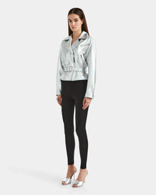A woman with long straight hair and a neutral expression stands against a plain white background, wearing the Rudsak ARIELLE - 8124052 Iridescent Cotton White jacket with black leggings and silver open-toe heeled sandals.