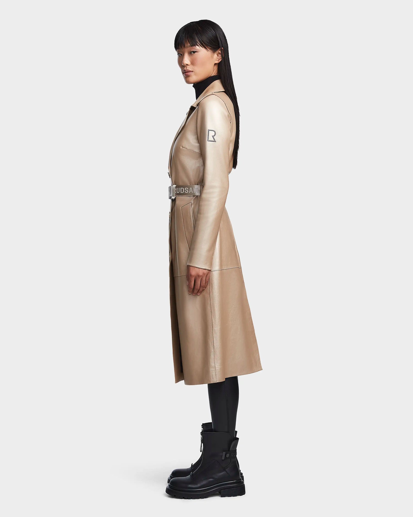 A woman with straight dark hair stands in profile, wearing the Rudsak KIRALY - 8123553 trench coat in metallic ivory, styled with black leggings and chunky black boots against a plain light background.