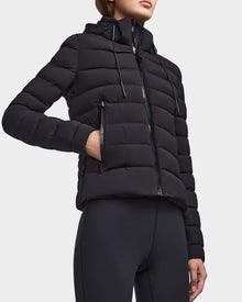Someone stands against a plain light background wearing the RUDSAK MELISSA - 8122551 BLACK fitted, weatherproof down jacket with a hood and zippered pockets, paired with black leggings.