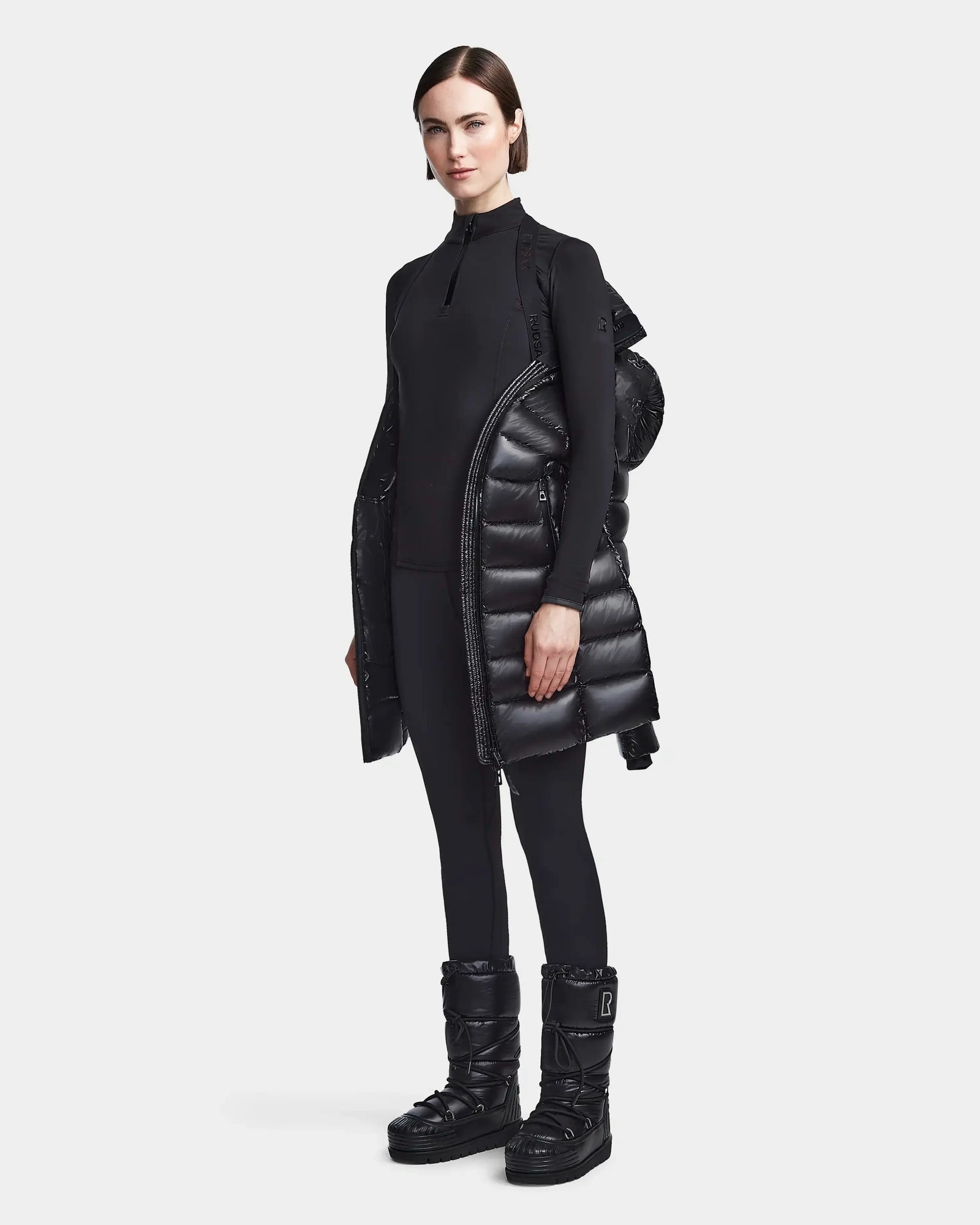 A woman stands against a white background wearing the Rudsak FRIDA S - L123513 Black women’s mid-length down jacket with removable hood, paired with a sleek black zip-up top, leggings, and black winter boots.