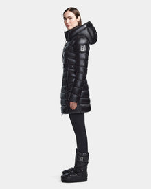 A woman stands in profile wearing the Rudsak FRIDA S - L123513 Black mid-length down jacket with a removable hood and belt, styled with black leggings and boots. The outfit features logo patches, set against a plain white background.