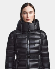 A woman with straight dark hair models the Rudsak FRIDA S - L123513 Black, a shiny black women's mid-length down jacket with a high hood and belted waist, standing against a plain white background.