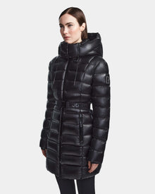 A woman stands slightly turned in a shiny black Rudsak FRIDA S - L123513 mid-length down jacket with large quilting, removable hood, and Rudsak branding on the belt and arm. The background is plain white.