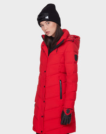 A woman in the Rudsak ELISA - L121512 POPPY RED quilted down puffer, with black gloves and a black knit beanie, stands against a plain white background, gazing slightly to the side.
