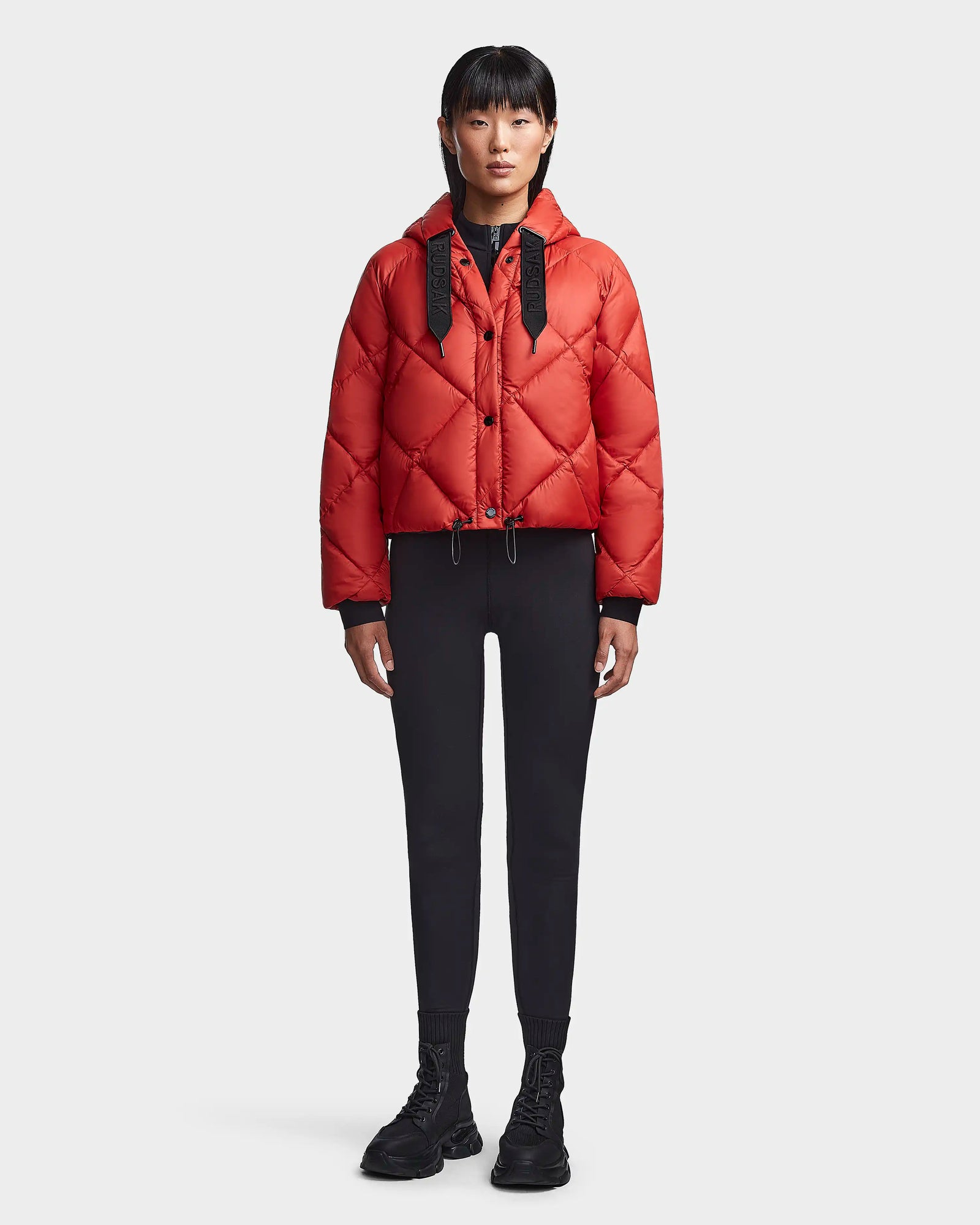 A person stands facing forward against a plain white background, wearing the Rudsak BALA - 8123614 Orange women’s winter coat with a hood, black drawstrings, black leggings, and chunky black shoes.